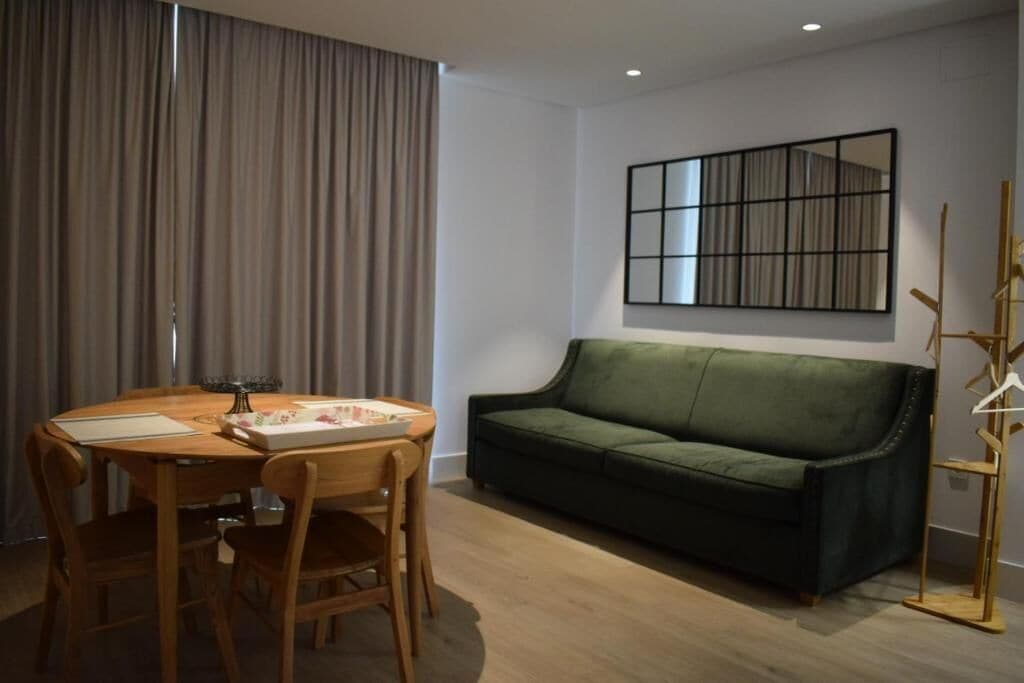 Una sala de estar moderna con una mesa de comedor de madera, un sofá verde y una decoración minimalista que incluye una obra de arte enmarcada y una lámpara de pie.