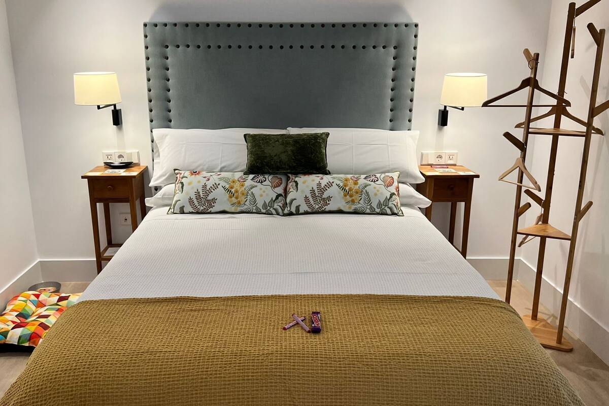 Ein ordentlich eingerichtetes Hotelzimmer mit einem großen Bett, zwei Nachttischen, einem hölzernen Kleiderhaken und einem farbenfrohen Kissen.