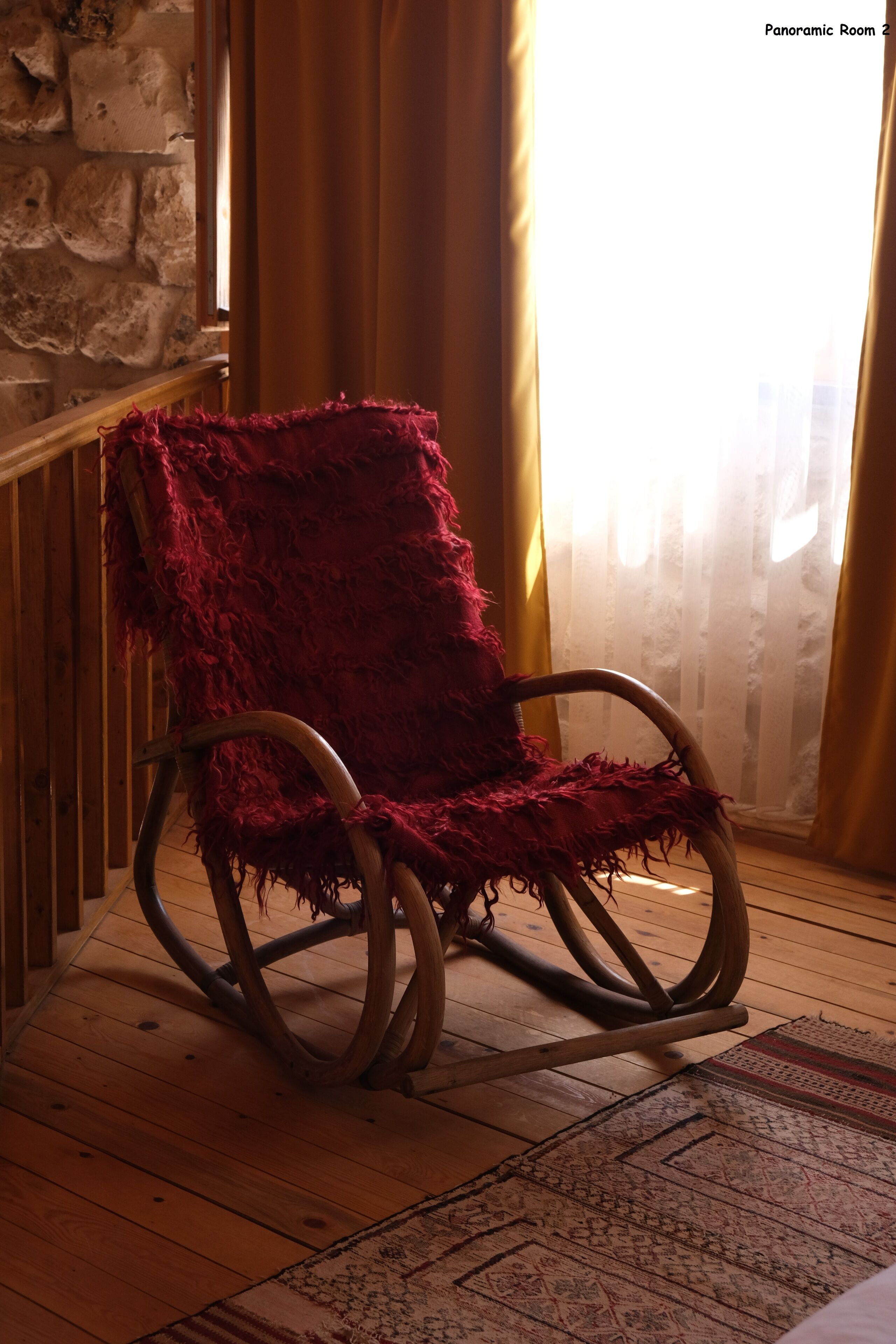 A rocking chair with a red blanket, wooden flooring, a patterned rug, and a stone wall.