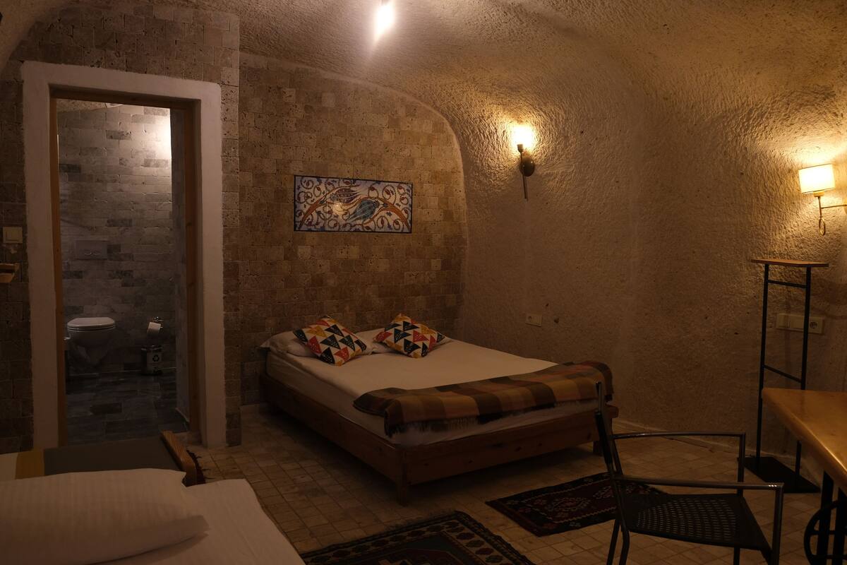 A bedroom with a bed, a chair, a table, a wall-mounted light, and a door leading to a bathroom.