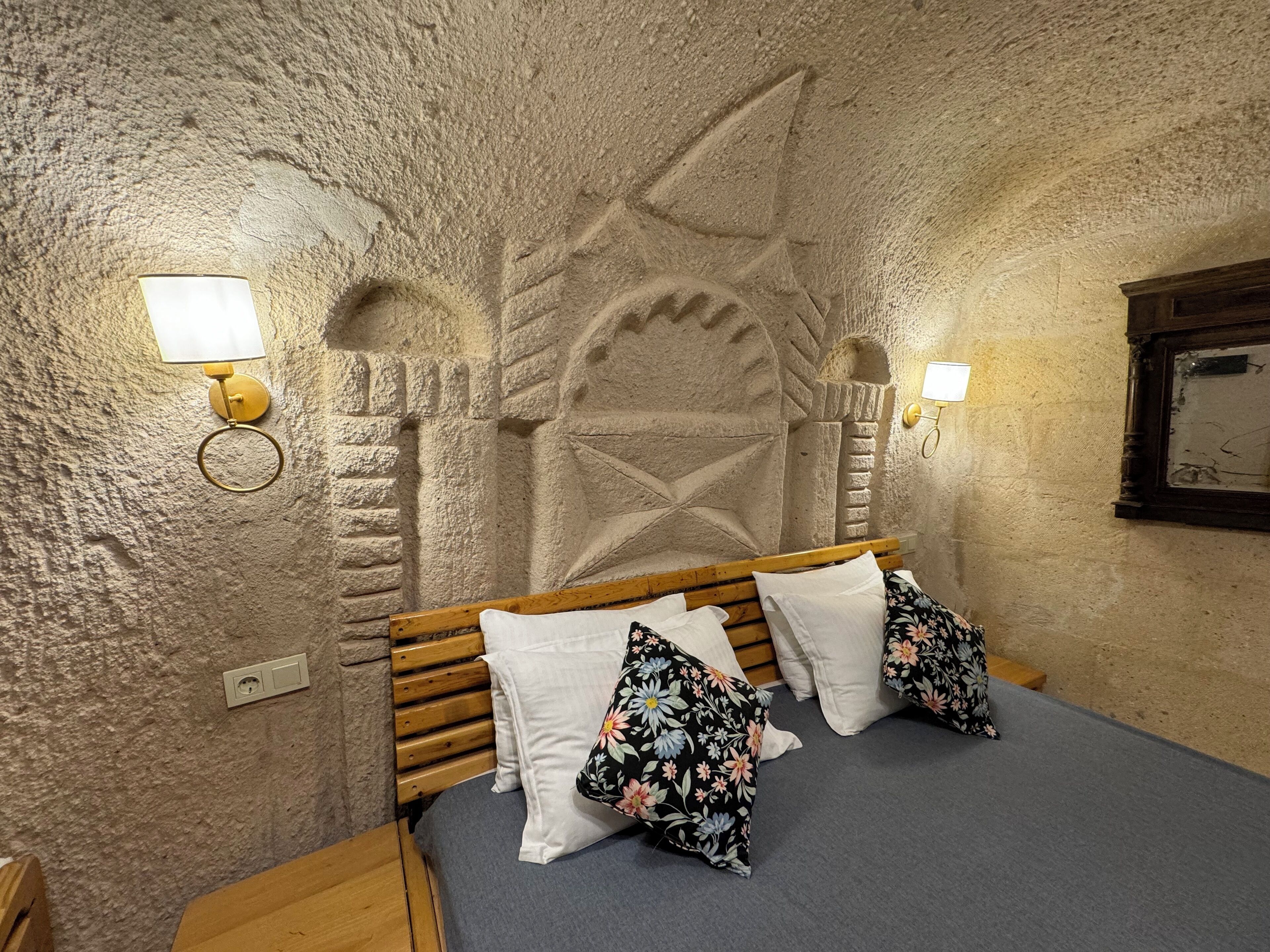 A room with a stone wall, a wooden bed with white and floral pillows, and wall-mounted lights.