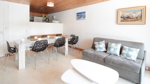 A modern living room with a wooden ceiling, a grey sofa with patterned cushions, a white dining table with chairs, and a kitchen area in the background. - Ski-In/Ski-Out 3-Bedroom Apartment, Sleeps 8, Flaine Forum, Stunning Mountain View (Flaine)