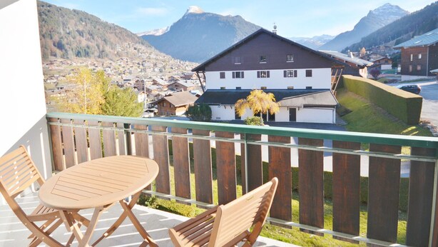 A balcony with a table and chairs, overlooking a mountain town with houses and a prominent mountain in the background.
