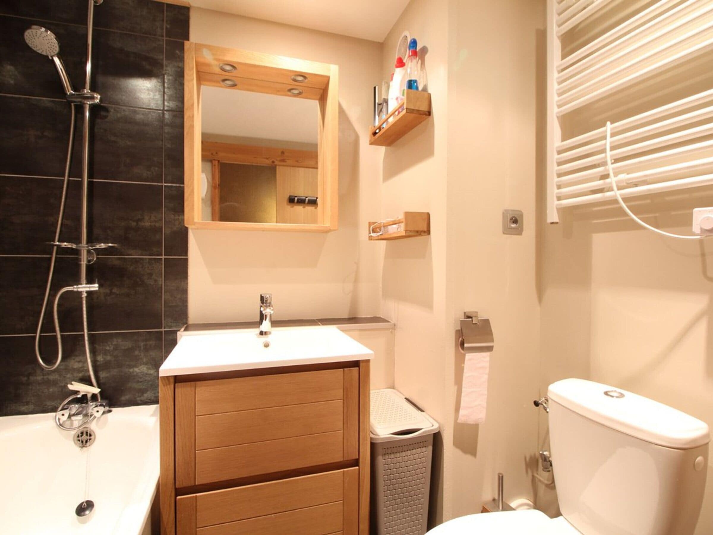 Un baño con un lavamanos blanco, tocador de madera, un espejo, ducha, inodoro y un perchero para toallas.