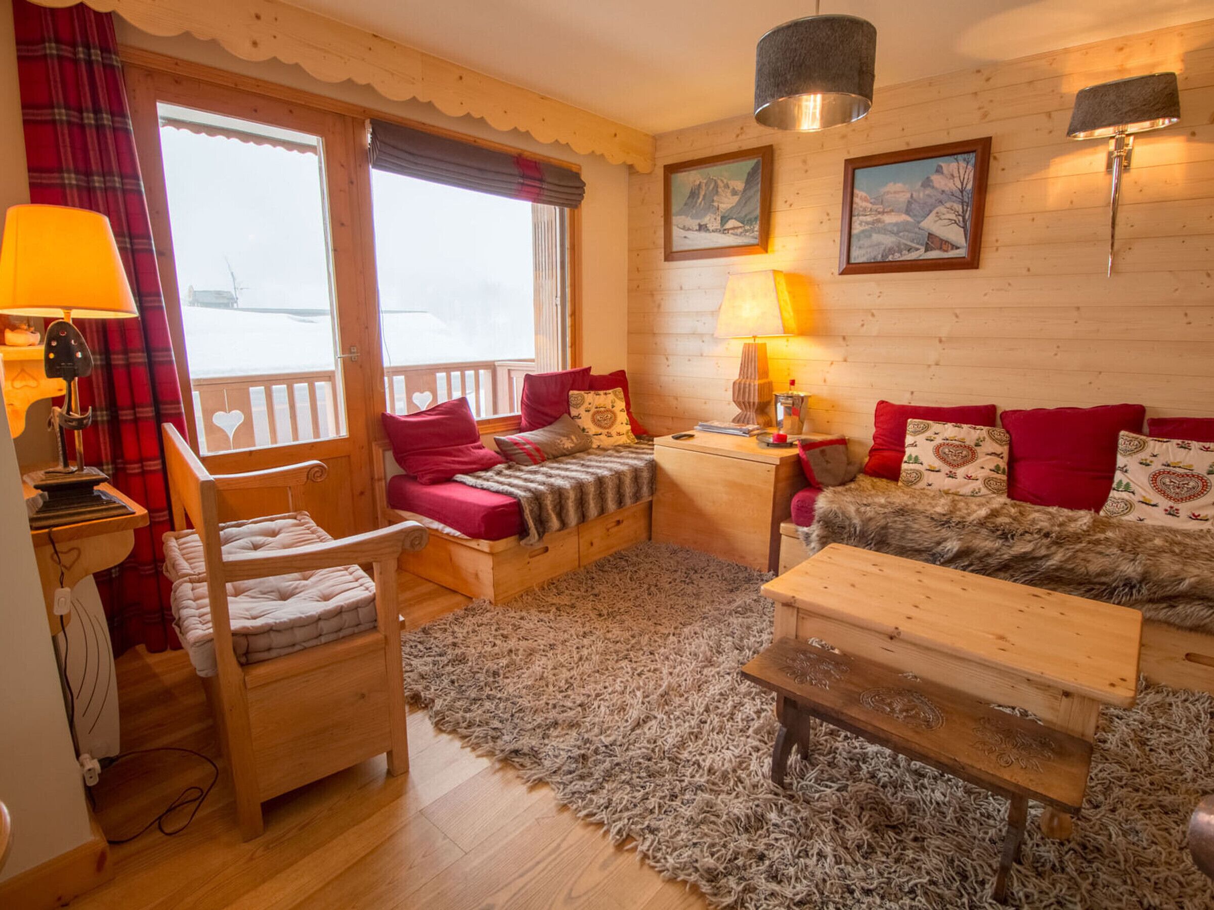 Ein gemütliches Zimmer mit einem hölzernen Bett, einem Holzstuhl, einem kleinen Tisch und einem Teppich. Es gibt zwei Fenster mit Vorhängen, eine Lampe auf einem Nachttisch und ein Bild an der Wand.