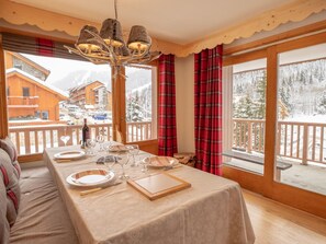 A cozy dining area with a table set for four, large windows with plaid curtains, and a view of a snowy mountain landscape. - Bright apartment in Méribel center with balcony and Wi-Fi (Méribel)