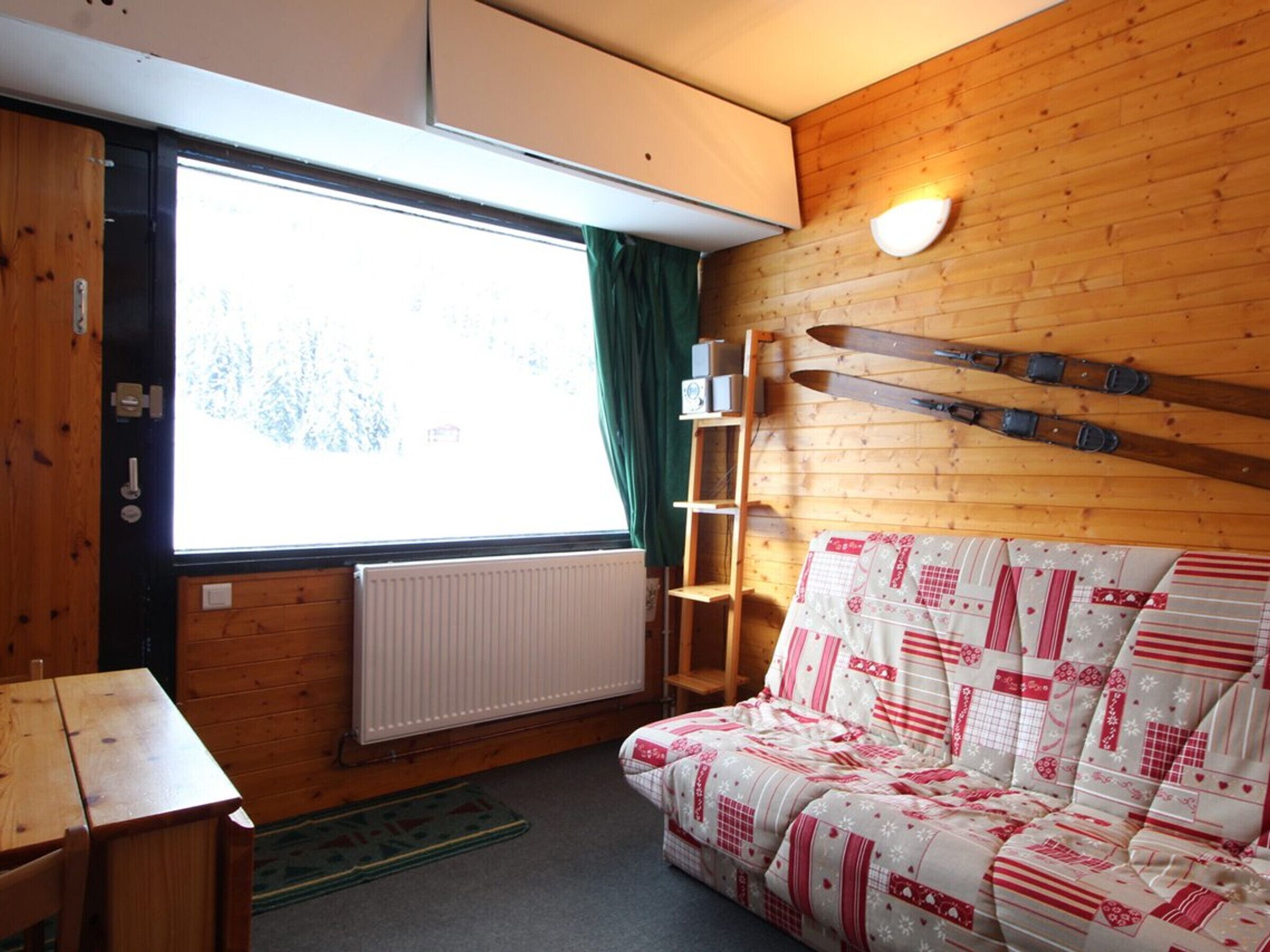 Una habitación de cabaña acogedora con interior de madera, un sofá con una manta estampada, una ventana con cortinas, un radiador y un par de esquíes apoyados en la pared.