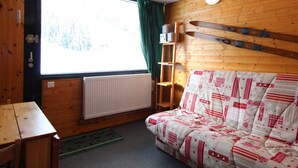 A cozy cabin room with a wooden interior, a sofa with a patterned blanket, a window with curtains, a radiator, and a pair of skis leaning against the wall.