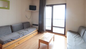 A compact living space with a sofa, a small table, a TV on a shelf, and a balcony with a view.