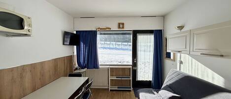 A compact room with a kitchenette, a sofa, a TV, and a window with curtains.