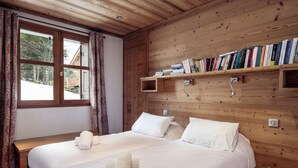 Een knus slaapkamer met twee bedden, een houten hoofdbord, een boekenplank vol boeken en een raam met gordijnen.