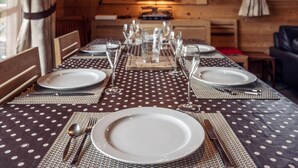 Een eettafel gedekt met borden, bestek en glazen, in een kamer met houten wanden en uitzicht op de keuken.
