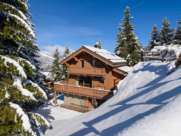 A wooden chalet with a balcony, surrounded by snow-covered trees and mountains.