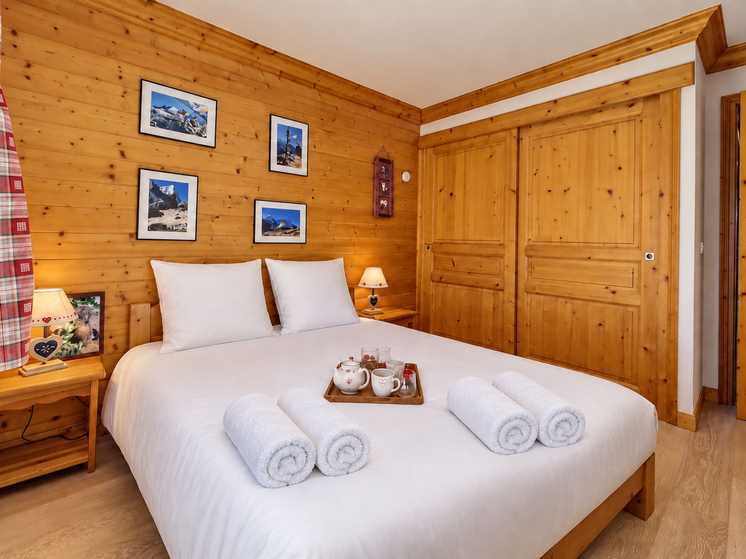 A wooden-paneled room with a bed, a nightstand, a tray with towels and a teapot, and framed pictures on the wall.