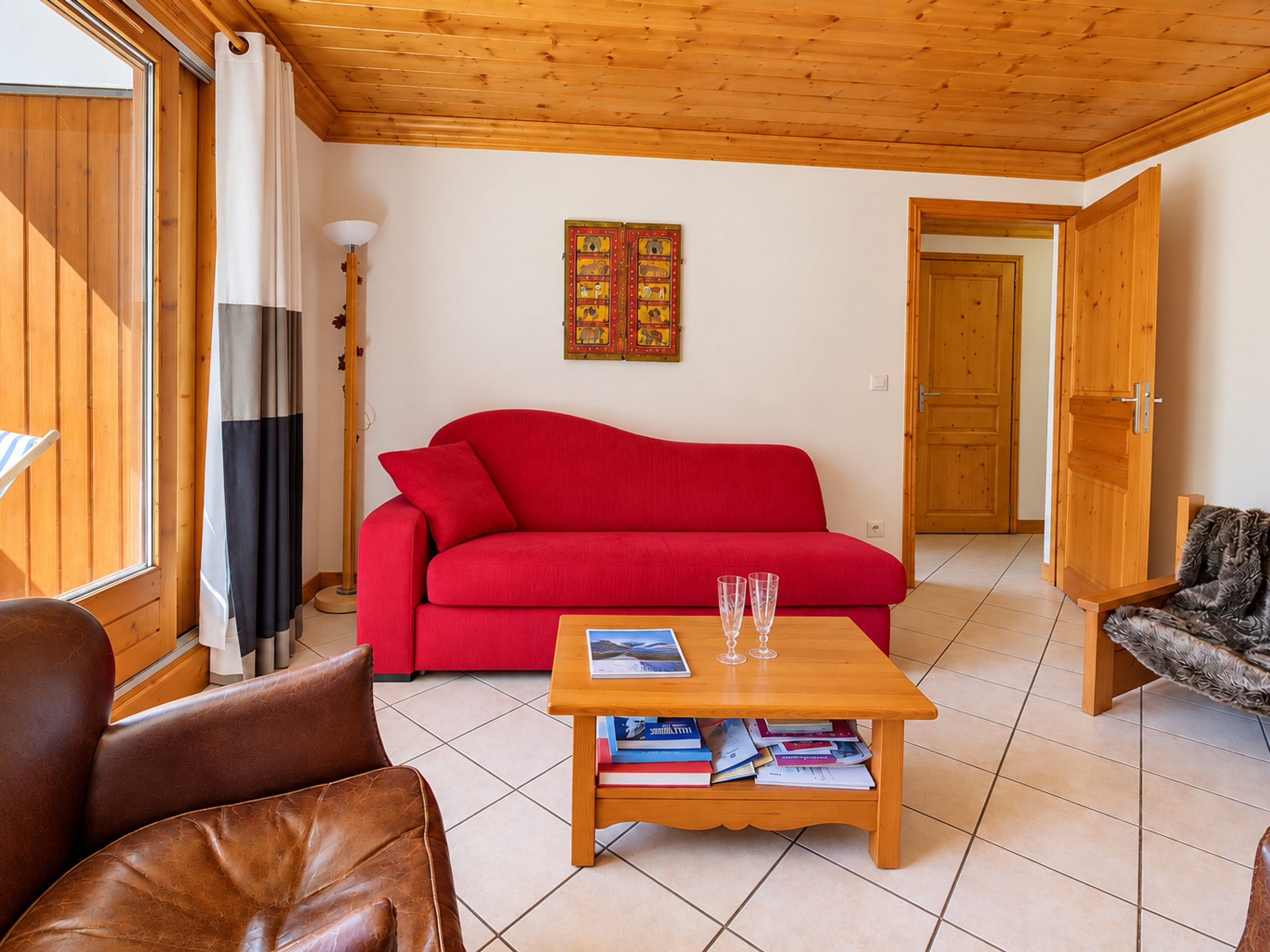 Ein Wohnzimmer mit einem roten Sofa, einem Holztisch mit Büchern und Gläsern, einer Lampe und einer Tür zu einem anderen Raum.