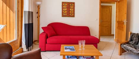 Ein Wohnzimmer mit einem roten Sofa, einem Holztisch mit Büchern und Gläsern, einer Lampe und einer Tür zu einem anderen Raum.