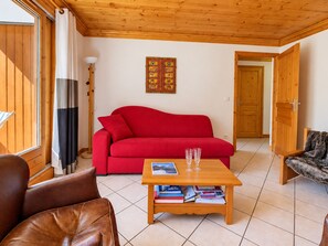 A living room with a red sofa, a wooden coffee table with books and glasses, a lamp, and a door leading to another room.