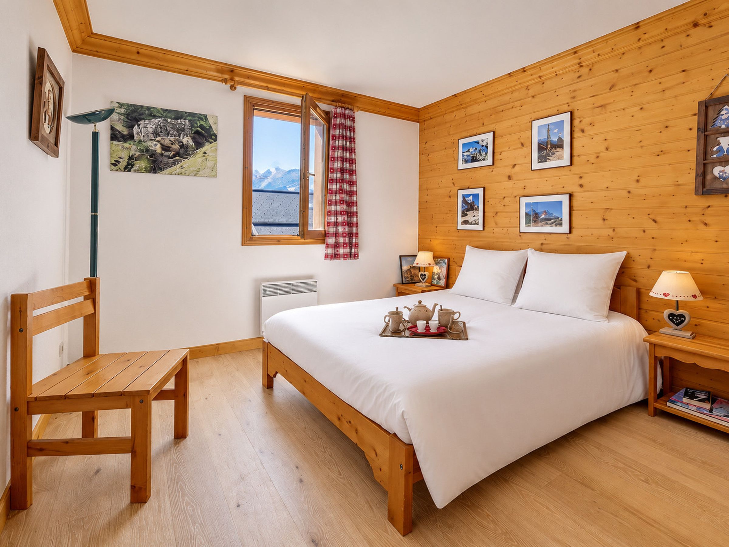 A bedroom with a wooden bed, a chair, a bedside table with a lamp, and a window with a view of mountains.