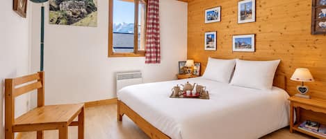 Un dormitorio con cama de madera, una silla, una mesita de noche con lámpara y una ventana con vistas a las montañas.
