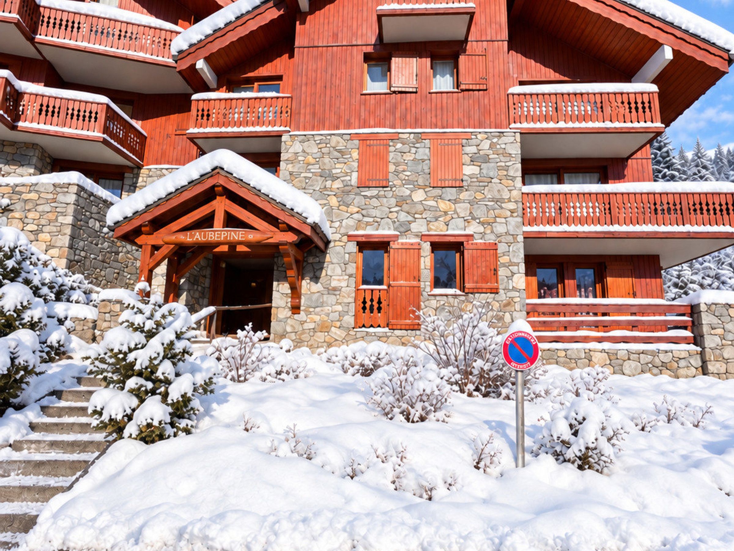 Un bâtiment en bois à plusieurs étages, doté de murs en pierre et de balcons. Une enseigne indique « L’AUBÉPINE », et un panneau « interdiction de stationner » est visible au premier plan.