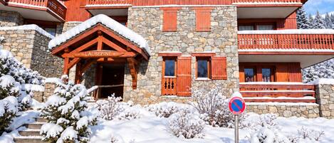 Un bâtiment en bois à plusieurs étages, doté de murs en pierre et de balcons. Une enseigne indique « L’AUBÉPINE », et un panneau « interdiction de stationner » est visible au premier plan.