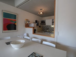 Una cocina moderna con armarios blancos, microondas y cafetera. Hay un comedor con una mesa y sillas blancas, y un tazón sobre la mesa.