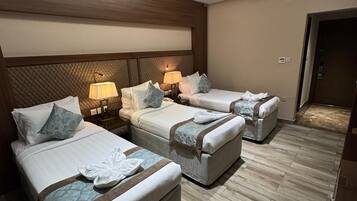 A hotel room with three beds, each with a pillow and a towel. There are two bedside tables with lamps. The room has a wooden headboard and a wooden floor.