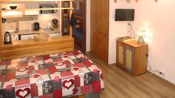 A compact kitchen with a microwave, kettle, and sink. A small table with a quilted tablecloth is in the center. A television is mounted on the wall, and a shelf with decorative items is above it.
