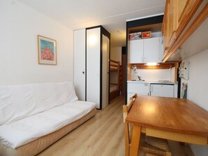 A compact living space with a white sofa, a wooden dining table, a kitchenette with cabinets, and a bunk bed in the background.