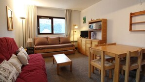 Ein gemütliches Wohnzimmer mit einem roten Sofa, einem Holztisch, einem Esstisch mit Stühlen, einem Fernseher auf einer Holzkommode und einem Fenster mit Vorhängen.