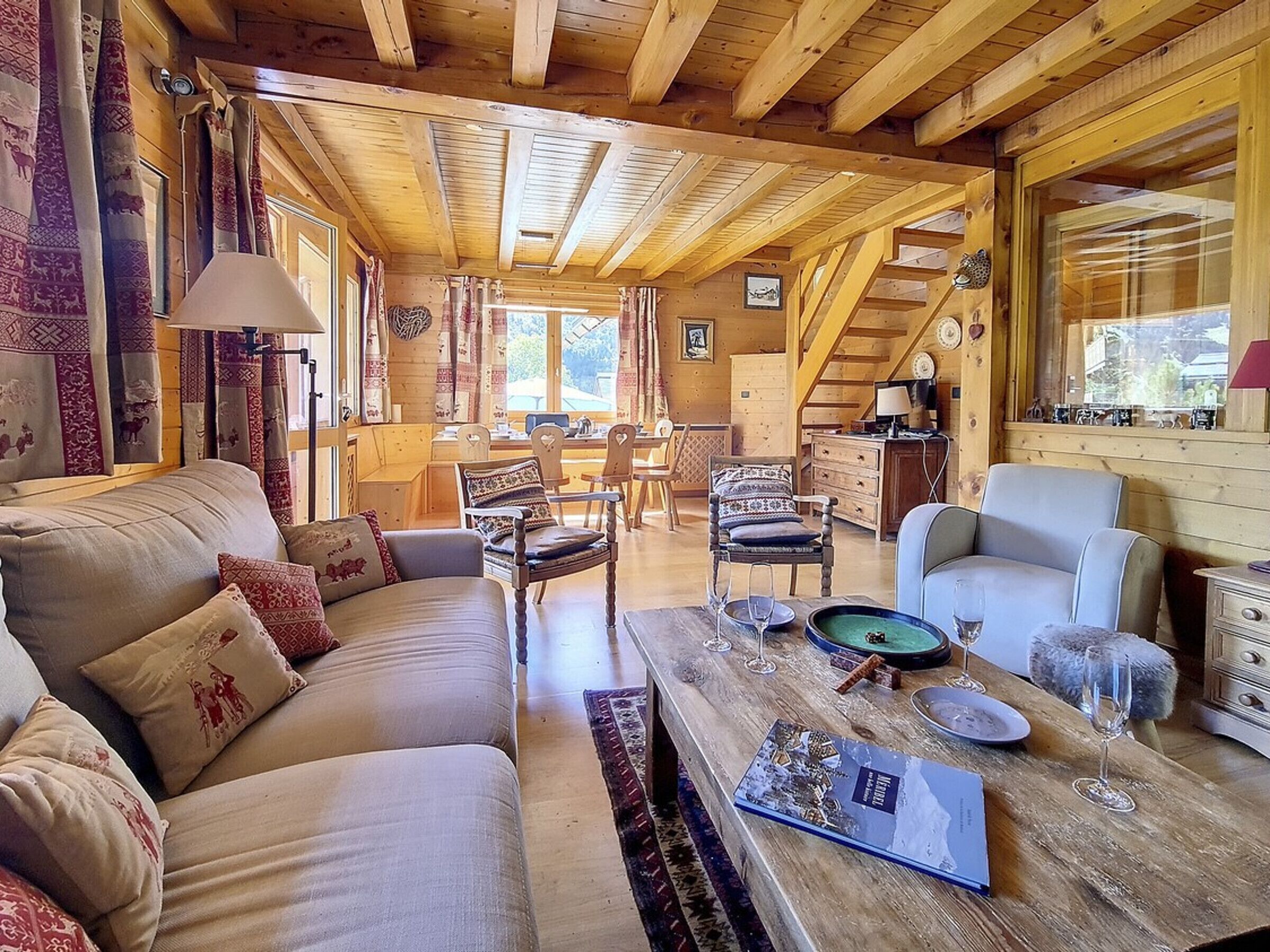 A cozy log cabin interior with a sofa, chairs, a dining table with wine glasses, and a staircase leading to an upper level.