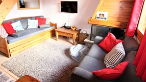 A cozy room with a wooden staircase, a sofa with red cushions, a grey couch, a small wooden table, and a television mounted on the wall.