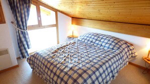 A bedroom with a bed, a window with curtains, a lamp, and a wooden ceiling.