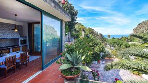 Eine moderne Villa mit großen Glasfenstern, die auf eine Terrasse führen, einem Essbereich mit Tisch und Stühlen sowie Blick auf das Meer und die Hügel.