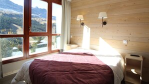 Een slaapkamer met een groot raam, houten wanden, een bed met een rode deken, twee bedlampen en een nachtkastje.