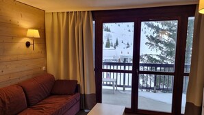 Een kamer met een houten wand, een rode bank, een klein tafeltje en uitzicht op een besneeuwd landschap met een houten terras.