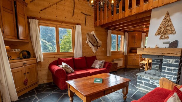 Interior acogedor de una cabaña con un sofá rojo, mesa de comedor de madera, suelo de piedra y chimenea.
