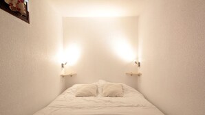 A narrow, white-walled room with a bed centered in the middle. There are two wall-mounted light fixtures on either side of the bed. - Cozy Studio for 5 in Sagittaire Residence, Near Slopes, South-Facing Balcony (Flaine)