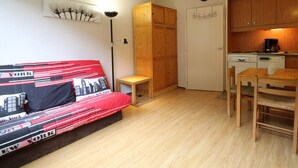 A compact room with a bed, a small table, a chair, a kitchenette, and a wardrobe.