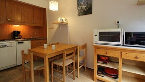 A compact kitchen with wooden cabinets, a dining table, chairs, a microwave, and a small shelf with kitchenware.