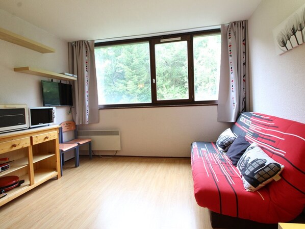 Une petite pièce agréable et chaleureuse, avec un bureau en bois, un canapé rouge, une fenêtre avec des rideaux et un téléviseur fixé au mur.