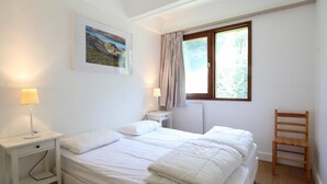 Een slaapkamer met een bed, nachtkastje, lamp, stoel, raam en een schilderij aan de muur.