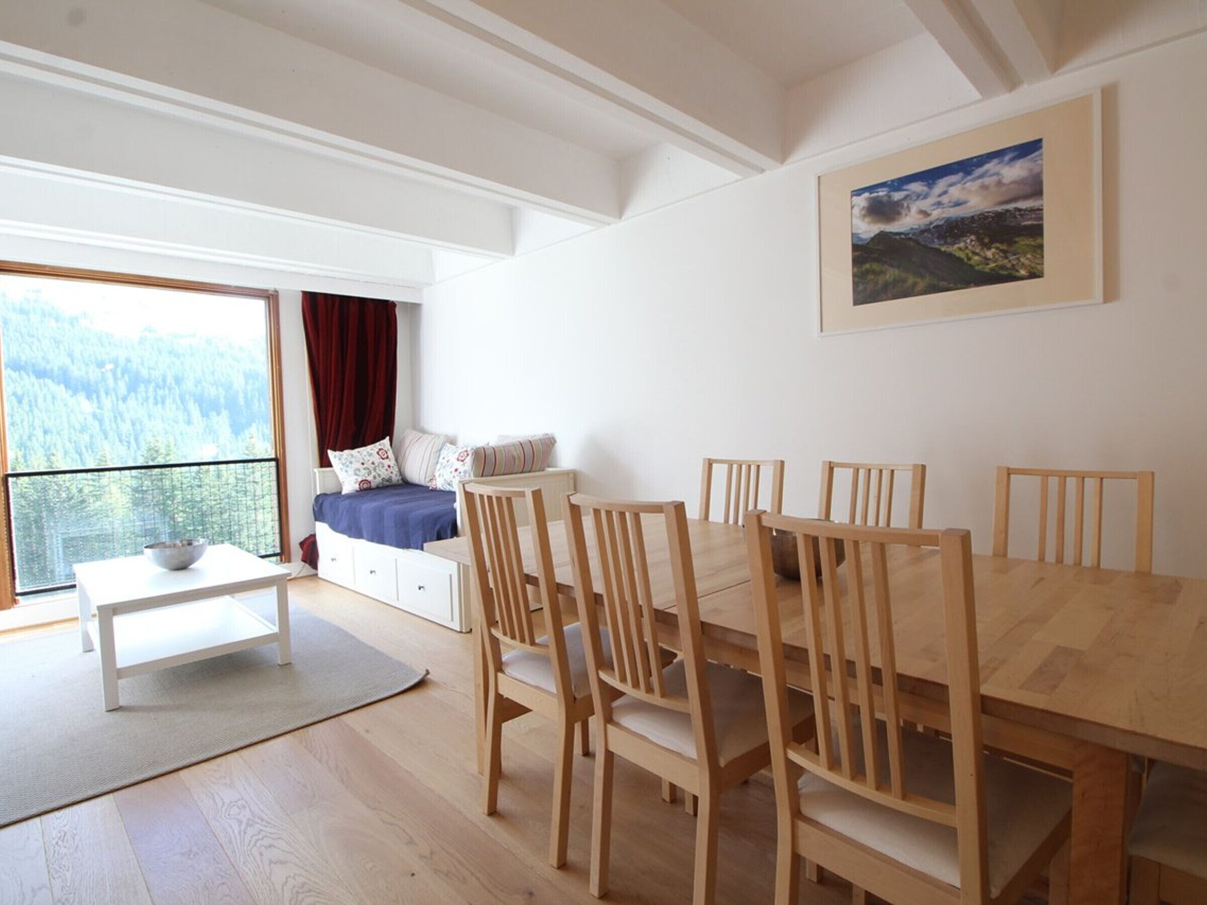 Una habitación con una mesa de comedor, sillas, un sofá con cojines, una mesita con un tazón y un ventanal con vistas a un bosque.
