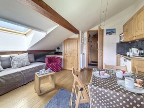Ein gemütlicher Loft-Raum mit Schlafgalerie, einem Esstisch für zwei Personen, einer kleinen Küchenzeile und einer Sitzecke mit Sofa und einem gemusterten Hocker.