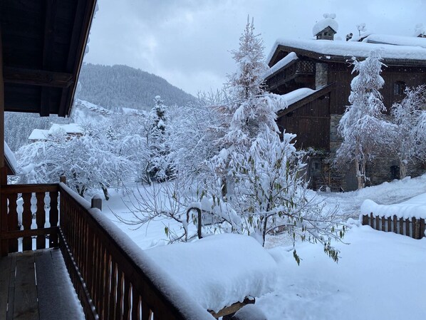 Un espace extérieur enneigé, doté d’une terrasse en bois, d’arbres chargés de neige et d’un bâtiment en pierre avec un toit en pente.