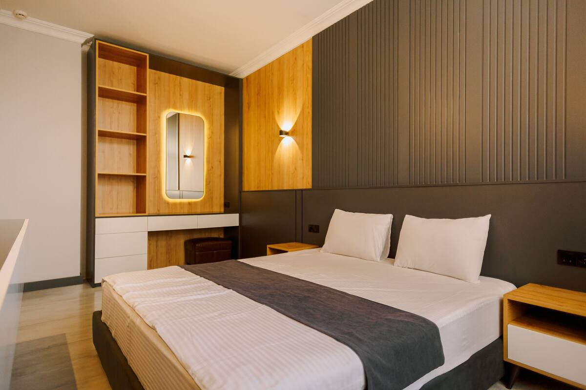 A modern hotel room with a large bed, a built-in wardrobe, a bedside table, and a wall-mounted light fixture.
