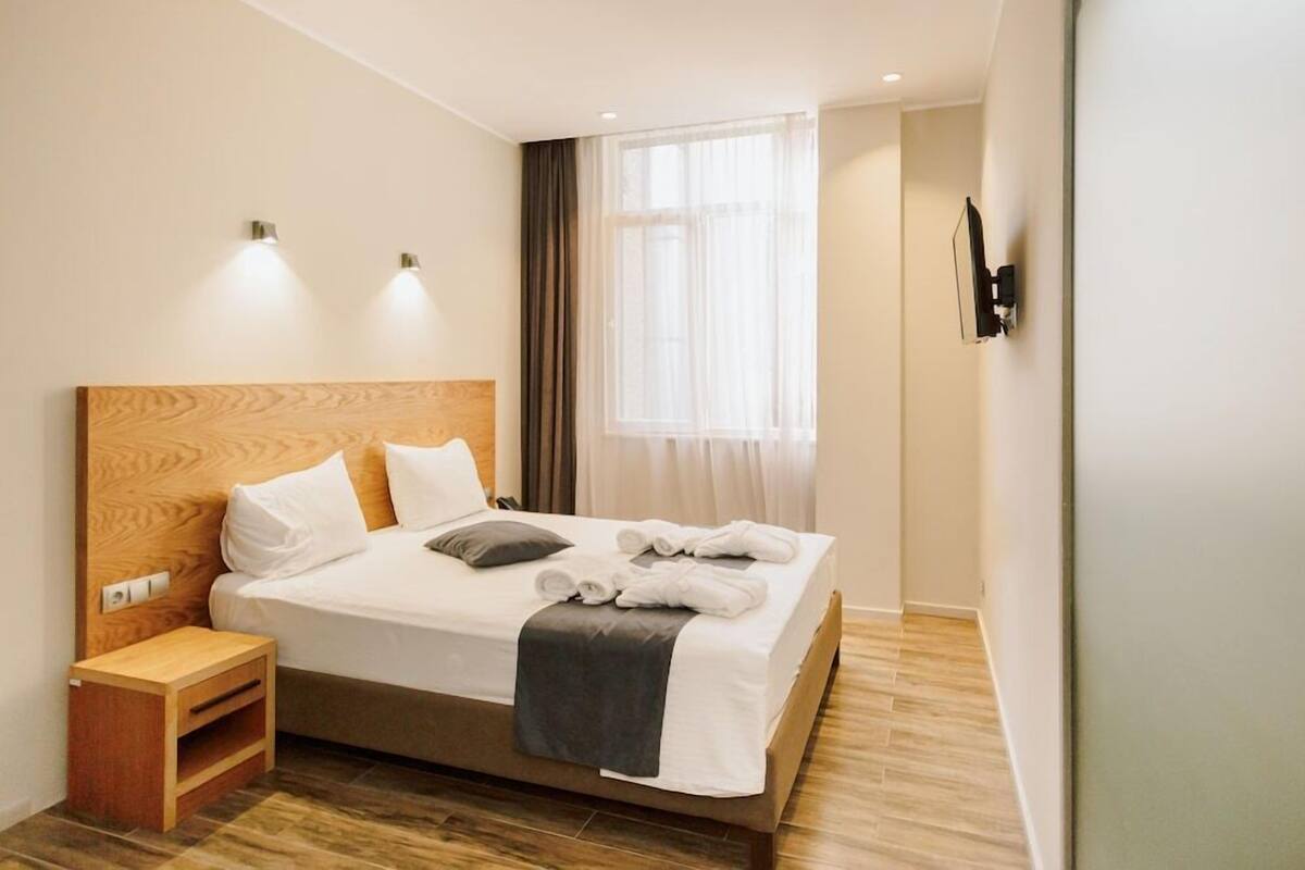 A hotel room with a wooden headboard, a bed with white linens, a bedside table, a TV mounted on the wall, and a window with sheer curtains.