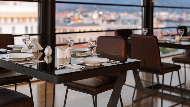 Una mesa de comedor con platos, vasos y cubiertos, con vistas a la ciudad a través de amplias ventanas.