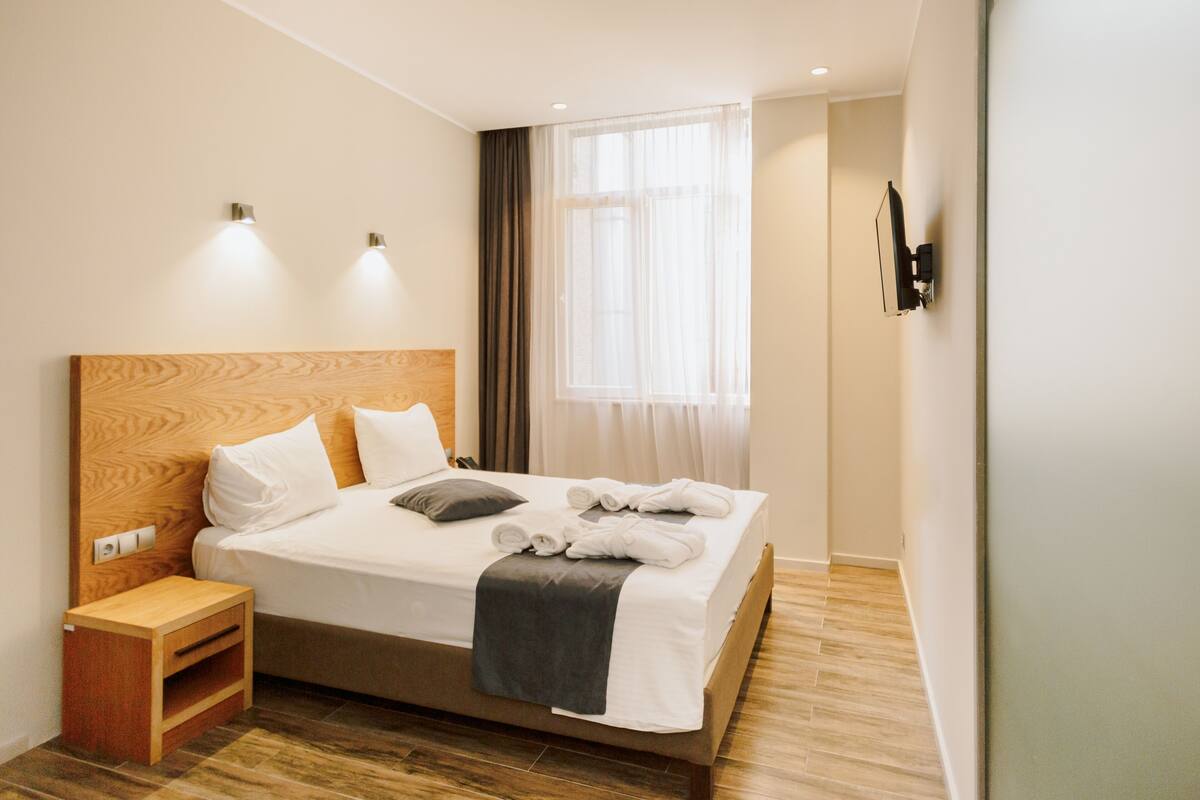 A neatly made bed with white linens and a gray throw blanket, a wooden nightstand with a drawer, and a wall-mounted TV.