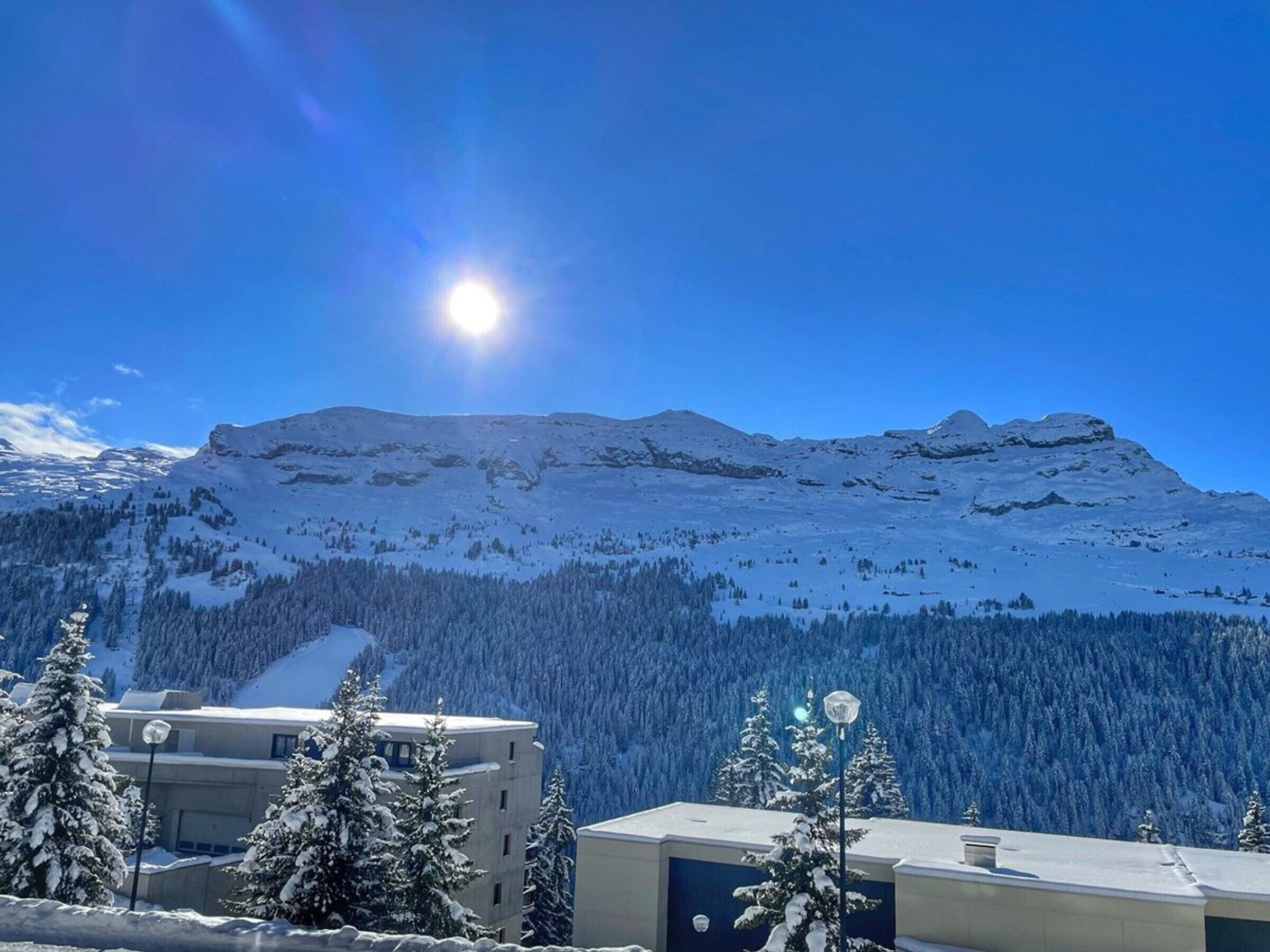 A snow-covered mountain with a clear sky and a bright sun. There are buildings with snow-covered roofs in the foreground.